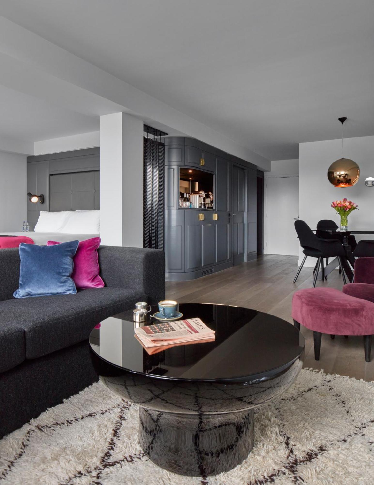 Hotel Sea Containers Londra Esterno foto The image shows a modern-looking living space, likely a hotel suite or stylish apartment. It features a dark gray sofa with colorful cushions in shades of blue and pink. In front of the sofa, there's a round black coffee table that holds a magazine a