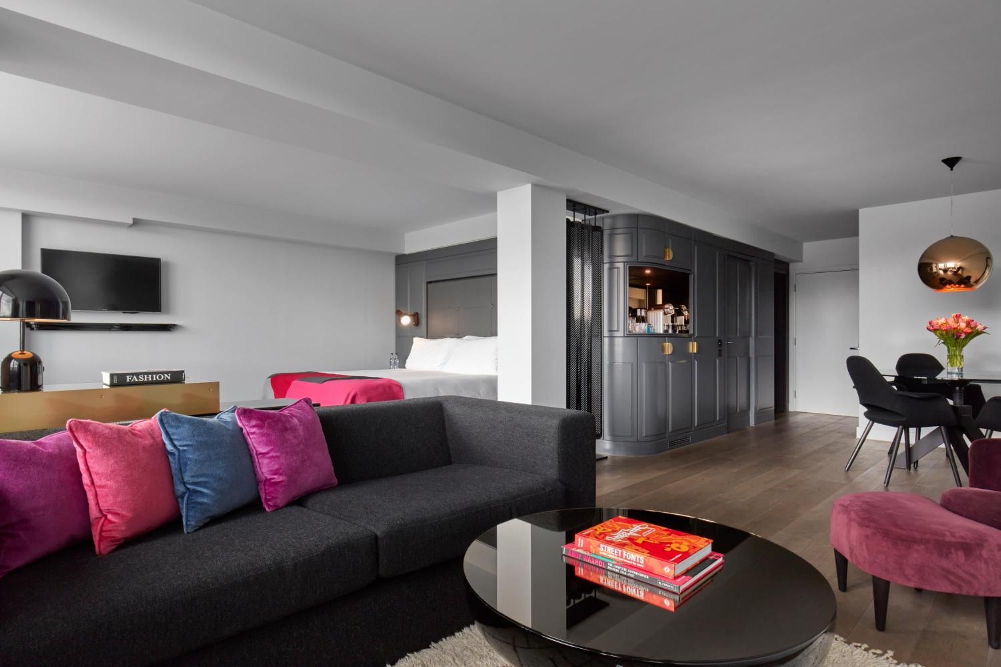 Hotel Sea Containers Londra Esterno foto The photo shows a modern and stylish living space. In the foreground, there is a dark gray sofa adorned with colorful pink and blue pillows. A circular coffee table sits in front of the sofa, with a few books stacked on it. 

In the background, there