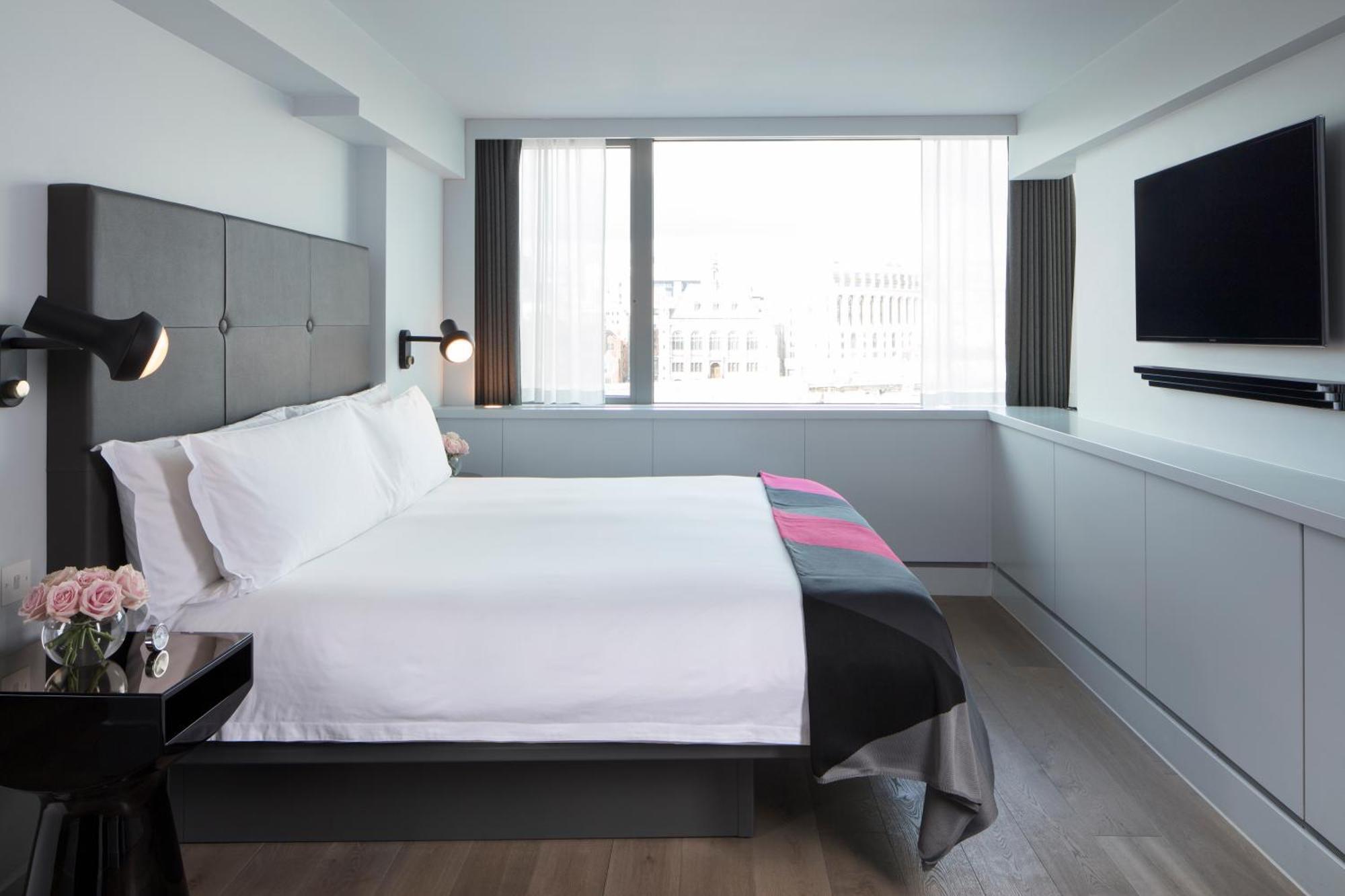 Hotel Sea Containers Londra Esterno foto The photo shows a modern hotel room with a minimalist design. There is a large bed with white linens, accompanied by a black and pink throw blanket at the foot. To the left of the bed, there is a small nightstand featuring a lamp and a bouquet of flo