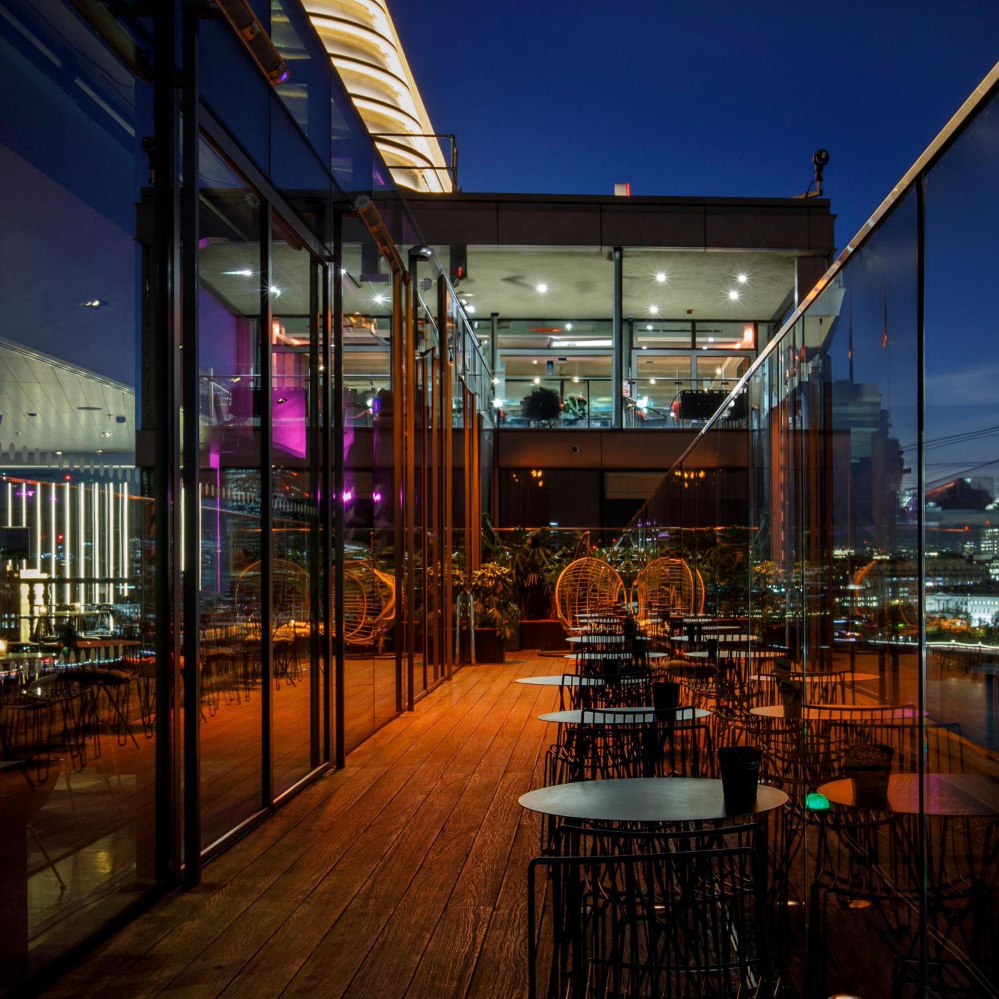 Hotel Sea Containers Londra Esterno foto The photo depicts a stylish restaurant or lounge area, featuring a modern design with extensive glass walls. It appears to be set on an upper floor, providing a view of the city skyline. The space has a wooden deck and several circular tables surroun