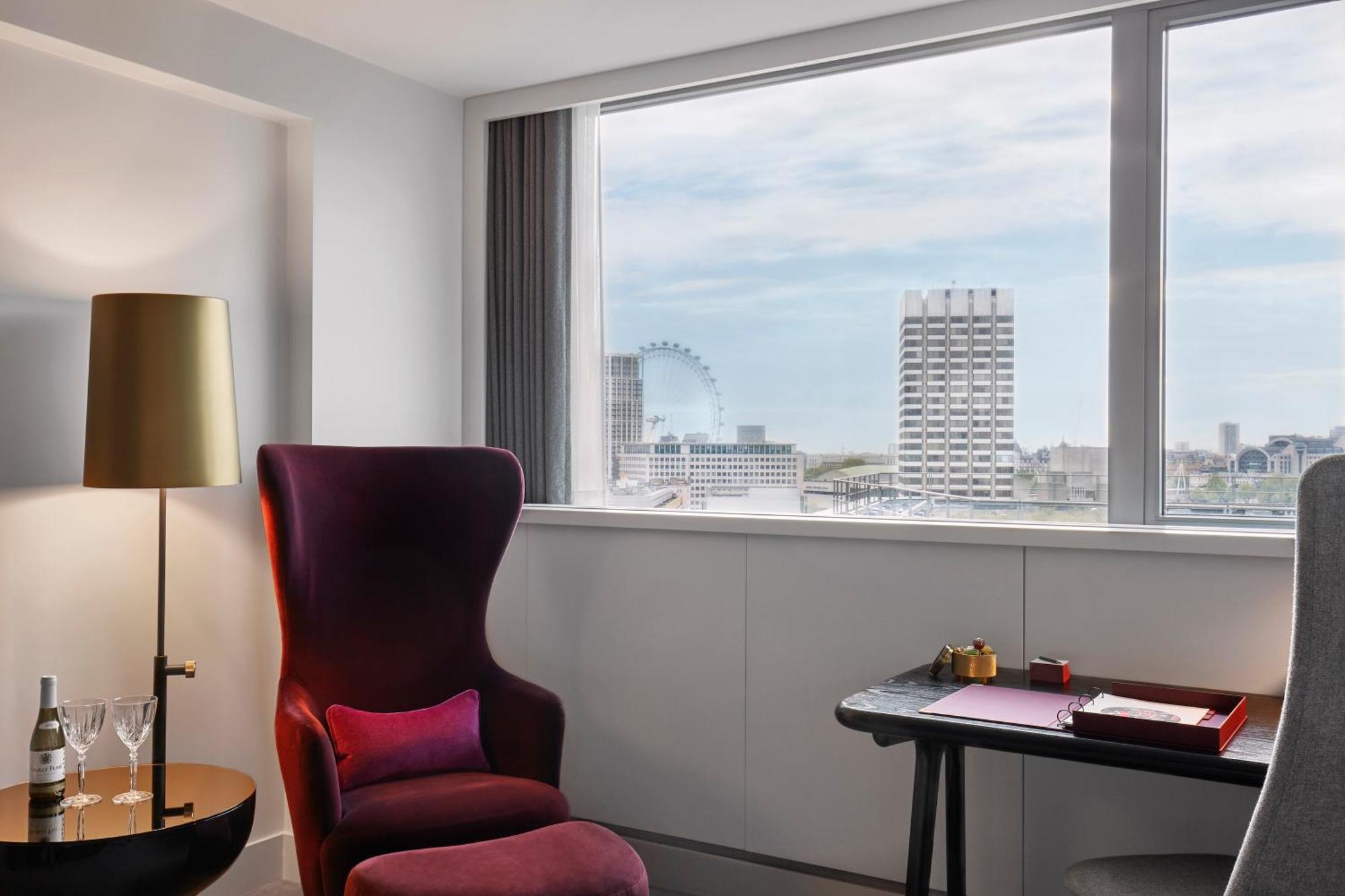 Hotel Sea Containers Londra Esterno foto The photo shows a modern and stylish hotel room featuring a large window with a view of the city skyline. There is a plush, deep purple armchair with a pink cushion in one corner, creating a cozy seating area. Next to the chair, there's a sleek floor