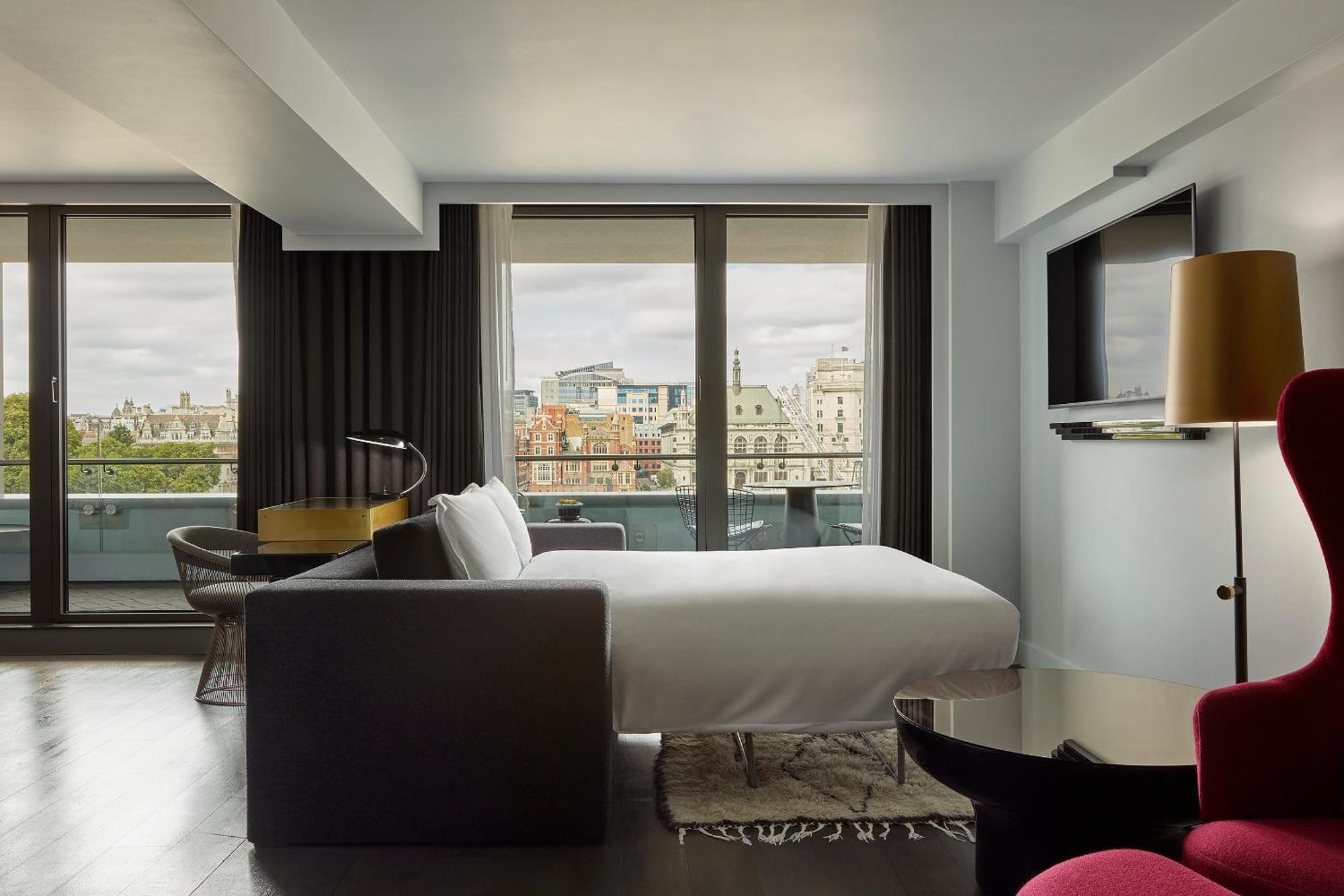 Hotel Sea Containers Londra Esterno foto The photo shows a modern hotel room with a large window offering a view of a cityscape. The room features a neatly made bed with a white duvet, a stylish gray sofa, and a small round table. There’s a flat-screen television mounted on the wall and a l