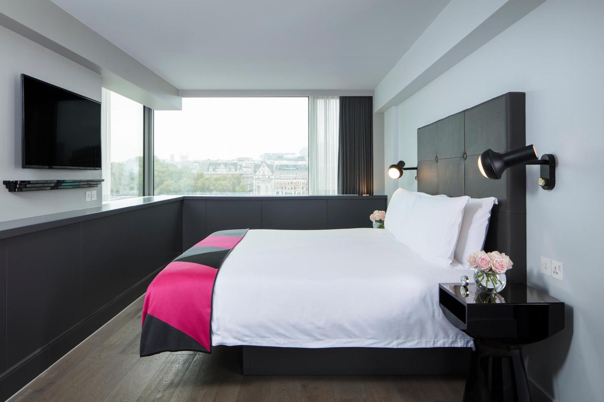 Hotel Sea Containers Londra Esterno foto The photo shows a modern hotel room featuring a large bed with a white comforter and a decorative blanket with pink accents. The headboard is black, and there are stylish wall-mounted lights on either side of the bed. A small bedside table with a vas
