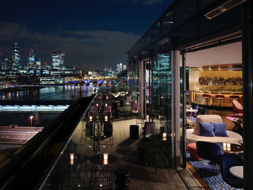 Hotel Sea Containers Londra Esterno foto The photo depicts a stylish rooftop bar or lounge area at night. There are modern seating arrangements both inside and on the outdoor terrace, which offers a scenic view of a city skyline across the river. The atmosphere is elegant, with soft lightin