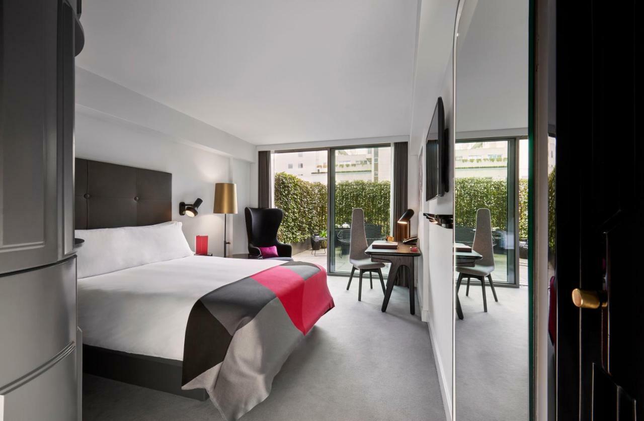 Hotel Sea Containers Londra Esterno foto The photo shows a modern hotel room featuring a large bed with a minimalist design. The bed is adorned with a colorful bedspread, primarily in shades of gray, white, and pink. There are two large windows that provide natural light and overlook a gree