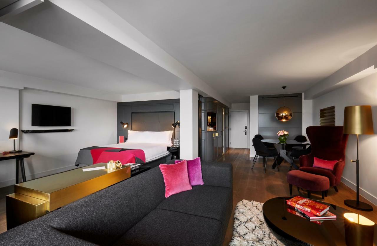 Hotel Sea Containers Londra Esterno foto The photo shows a modern hotel room that features a stylish design. On one side, there is a gray couch with vibrant pink pillows, complemented by a sleek glass coffee table. Across from the couch, a bed with white linens is visible, set against a dar