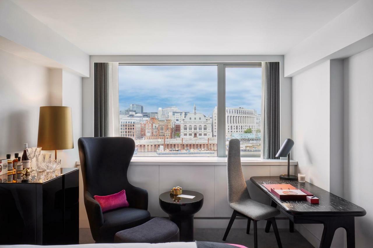 Hotel Sea Containers Londra Esterno foto The photo shows a modern hotel room with a stylish aesthetic. There is a large window that offers a view of the city skyline. The room features a comfortable armchair in dark upholstery with a pink accent pillow, a small round table, and a sleek desk