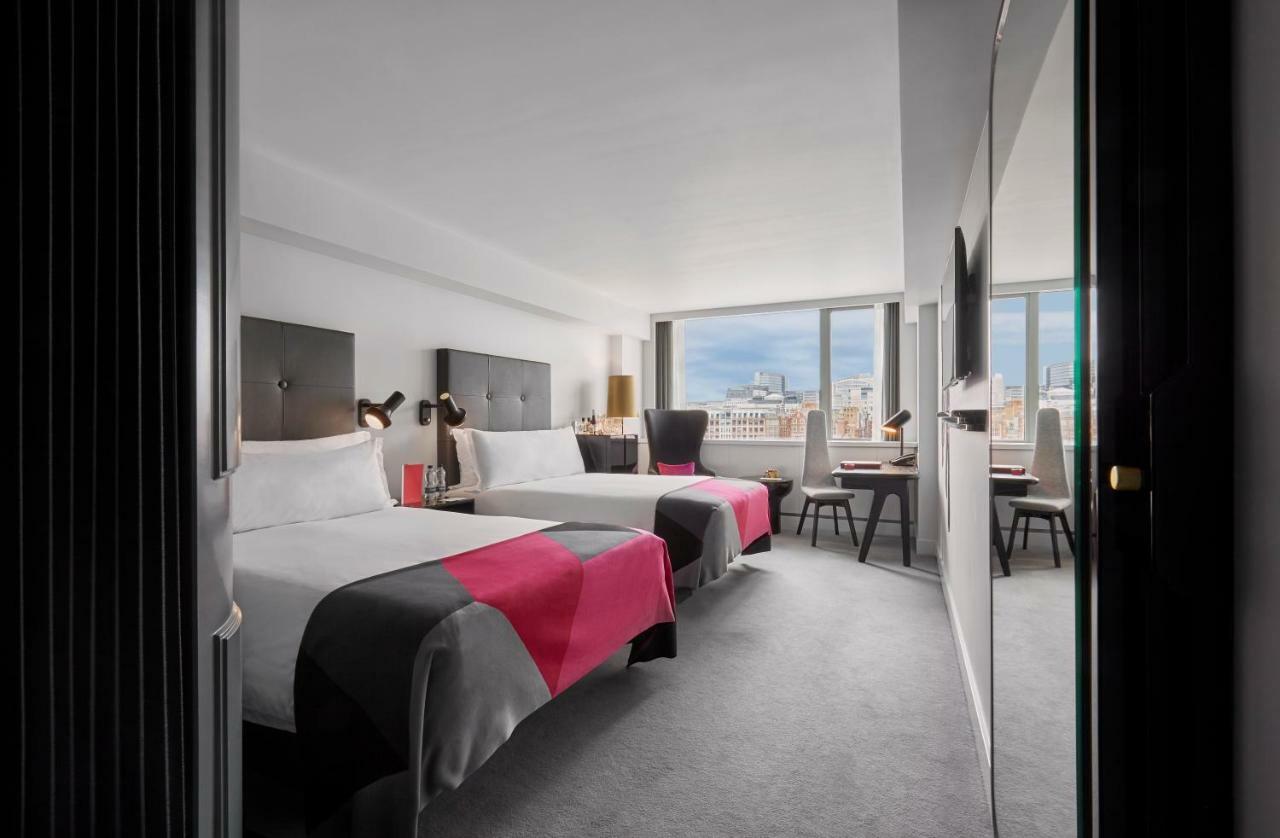Hotel Sea Containers Londra Esterno foto The photo shows a modern hotel room featuring two beds with stylish bedding, largely in gray and pink colors. The room is well-lit, with large windows that provide a view of the outside landscape. There are also some pieces of furniture, including a 