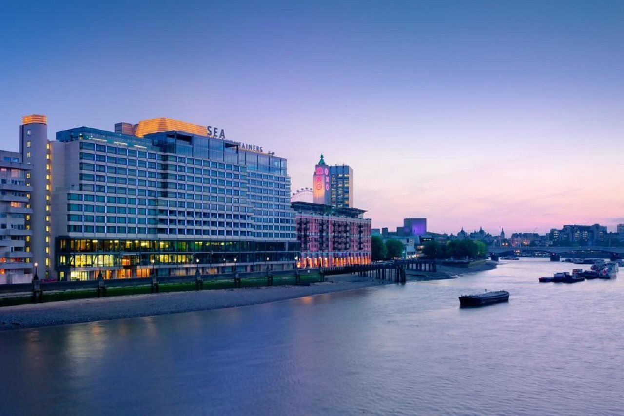Hotel Sea Containers Londra Esterno foto The photo shows a waterfront scene featuring a modern hotel or building along a river. The structure has a large façade with lights illuminating it, suggesting it is an upscale destination. The sky is transitioning from daylight to twilight, displayi