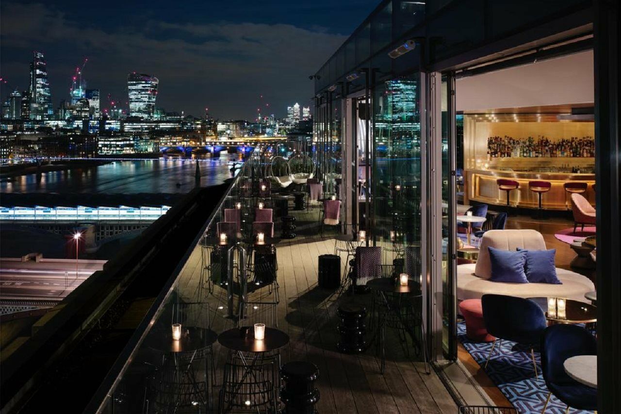 Hotel Sea Containers Londra Esterno foto The image showcases a modern rooftop bar or lounge with a stunning nighttime cityscape view. The terrace is lined with stylish seating arrangements, including lounge chairs and small tables, all adorned with candles that create a cozy atmosphere. In 